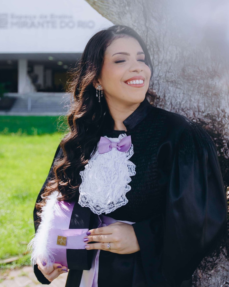 Formanda de beca fotografada ao ar livre, com cabelo e mãos visíveis